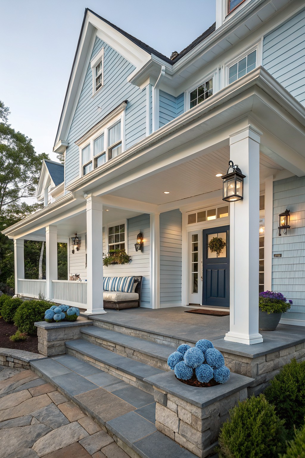A two-story light blue house with white trim, a dark blue front door, covered porch with white columns, and stone steps leading to the entry.