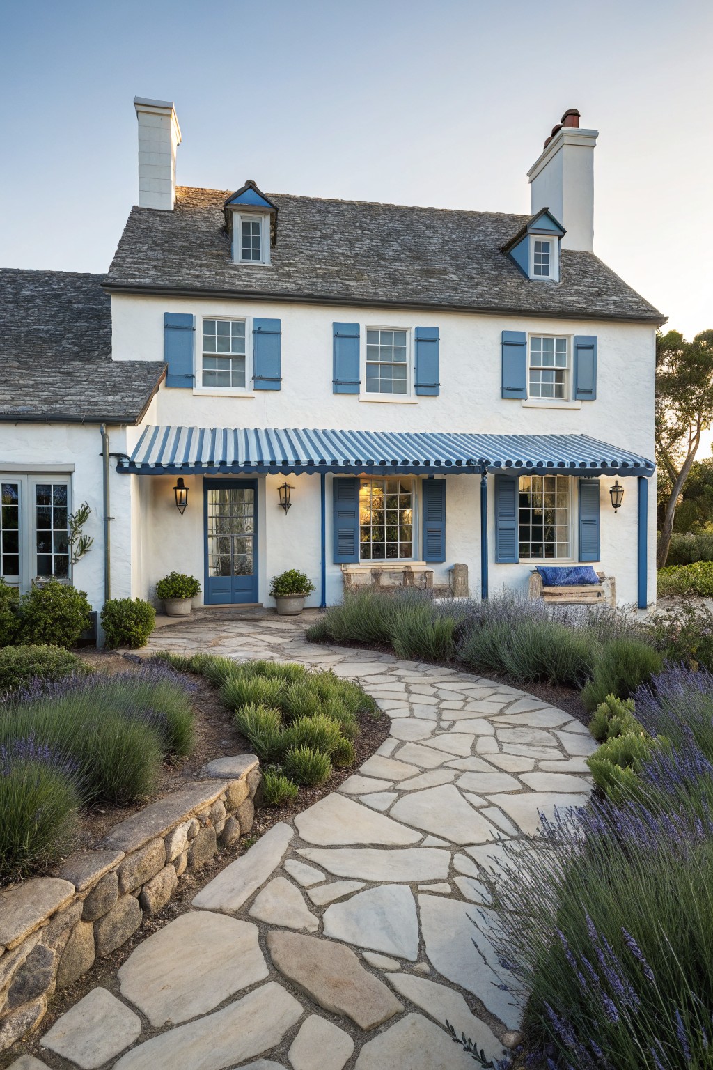 A white two-story house with blue shutters, blue door, and blue striped awning, approached by a curved stone path with lavender plantings.