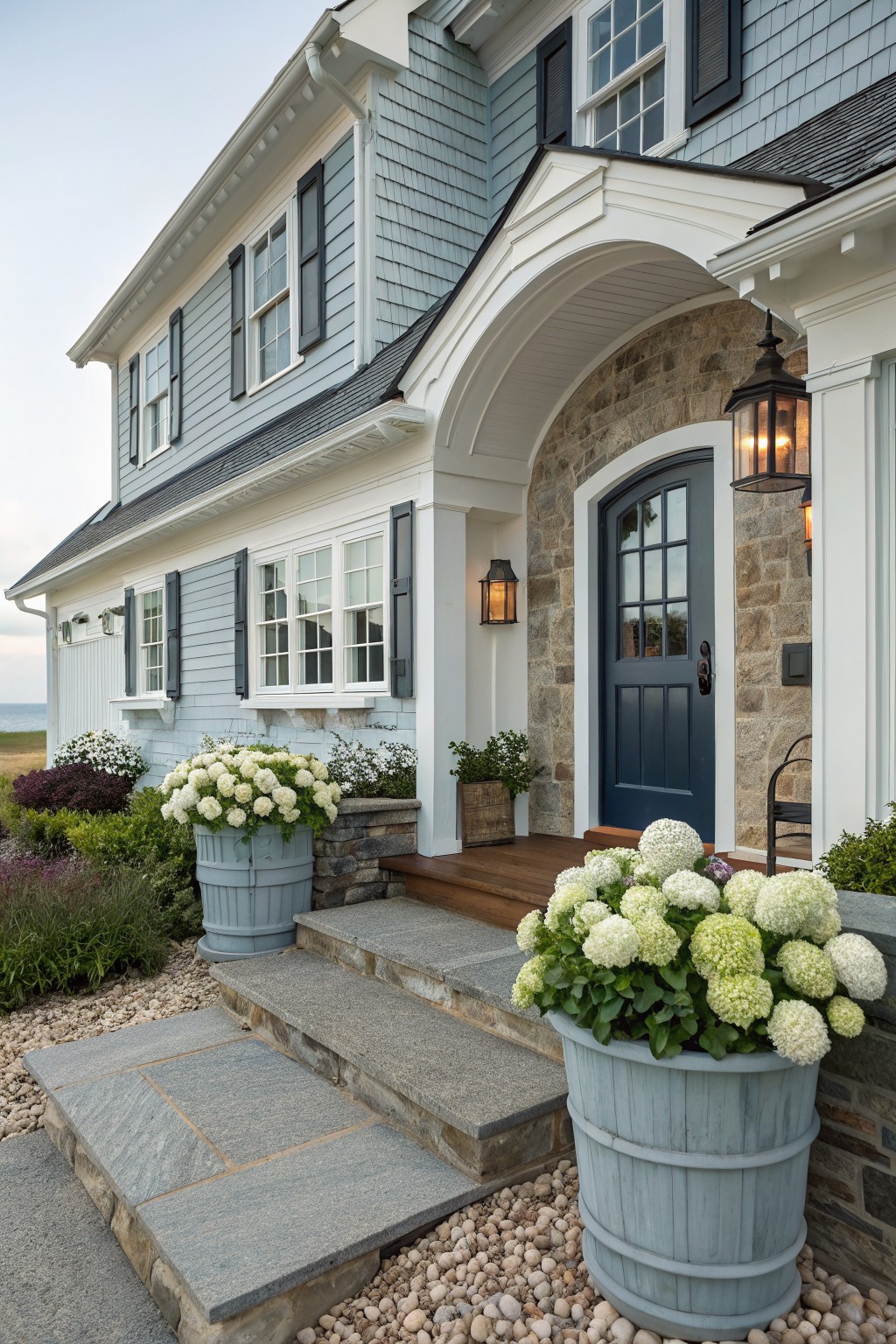 A two-story coastal house with light blue shingle siding, white trim, a dark blue front door set in stone, and large gray planters holding white flowers beside stone steps.