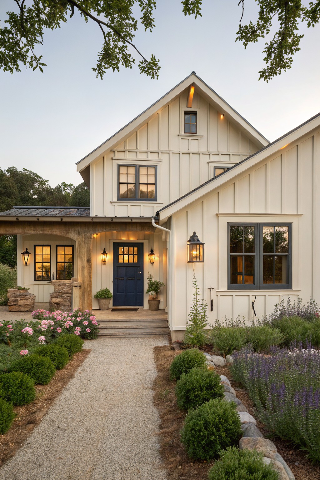 A white farmhouse with board and batten siding features a centered navy blue front door under a gable, flanked by windows and exterior lights, with a gravel path and garden beds leading to the entrance.