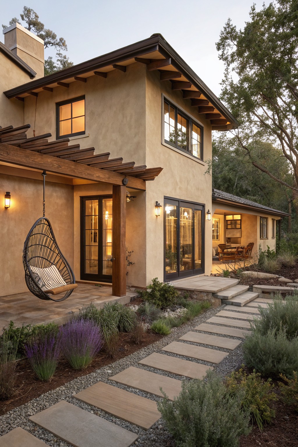 A stucco ranch house with dark wood trim, a covered entry featuring a hanging chair, and a stone pathway through landscaped beds.