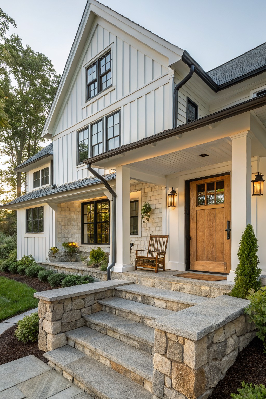 A white board and batten farmhouse exterior with a prominent front gable, stone foundation accents, a covered porch with white columns, and a wooden front door.