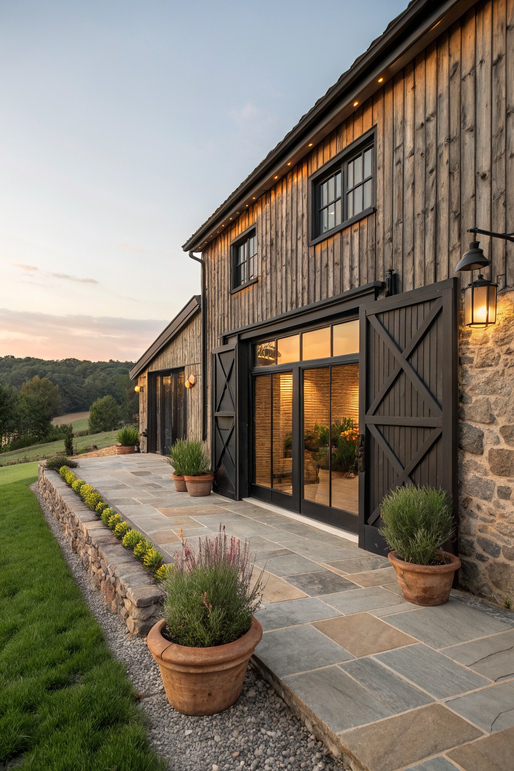 A wooden farmhouse exterior with vertical siding, large black barn doors, black-framed windows, and a stone patio lined with potted plants.