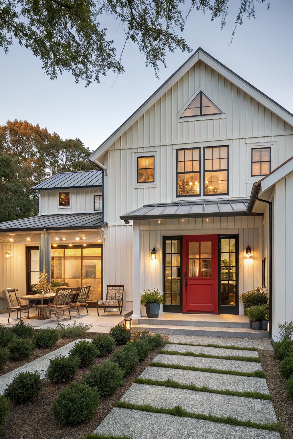 A white board and batten farmhouse with a red front door, black-framed windows, a metal roof overhang, and a stone pathway leading to the entrance.