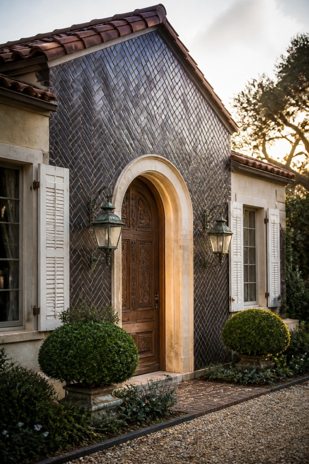 A house exterior with a wooden door under a stone arch, herringbone tile above the entry, and lanterns mounted on either side.