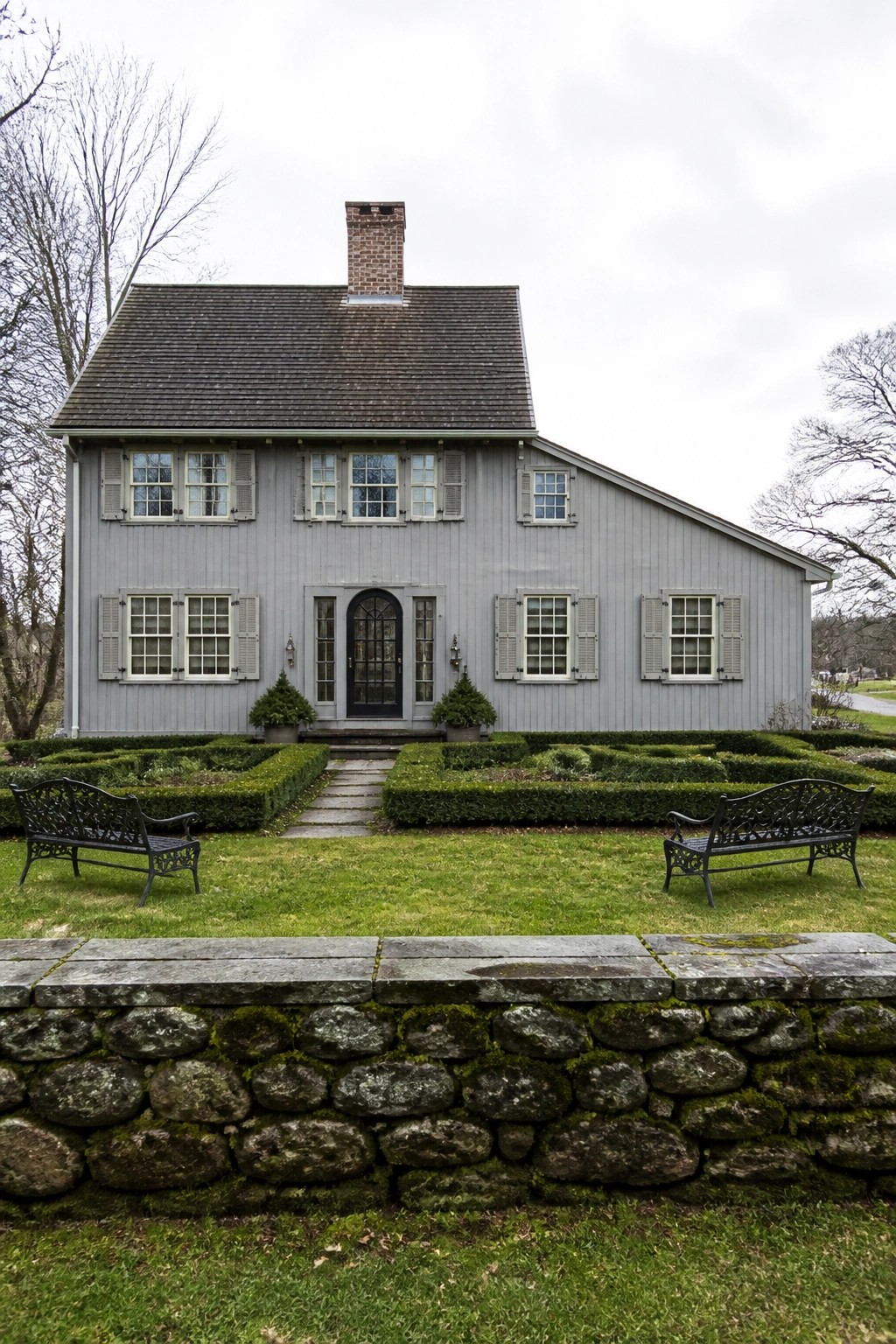 A gray two-story farmhouse with symmetrical windows flanking a central black door, set behind a low stone wall and boxwood garden.