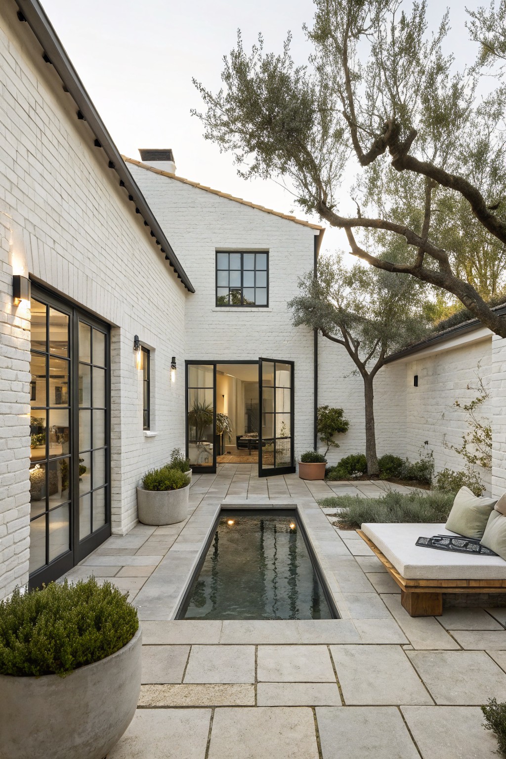 A white brick house exterior with black-framed glass doors opening onto a stone paver courtyard that includes a rectangular reflecting pool and scattered potted plants.