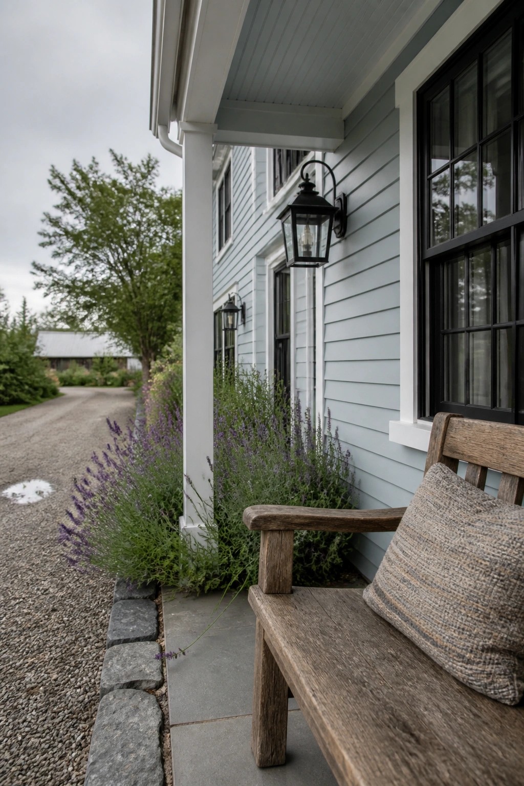 Wooden bench with pillow on blue house porch beside gravel path and flowers.