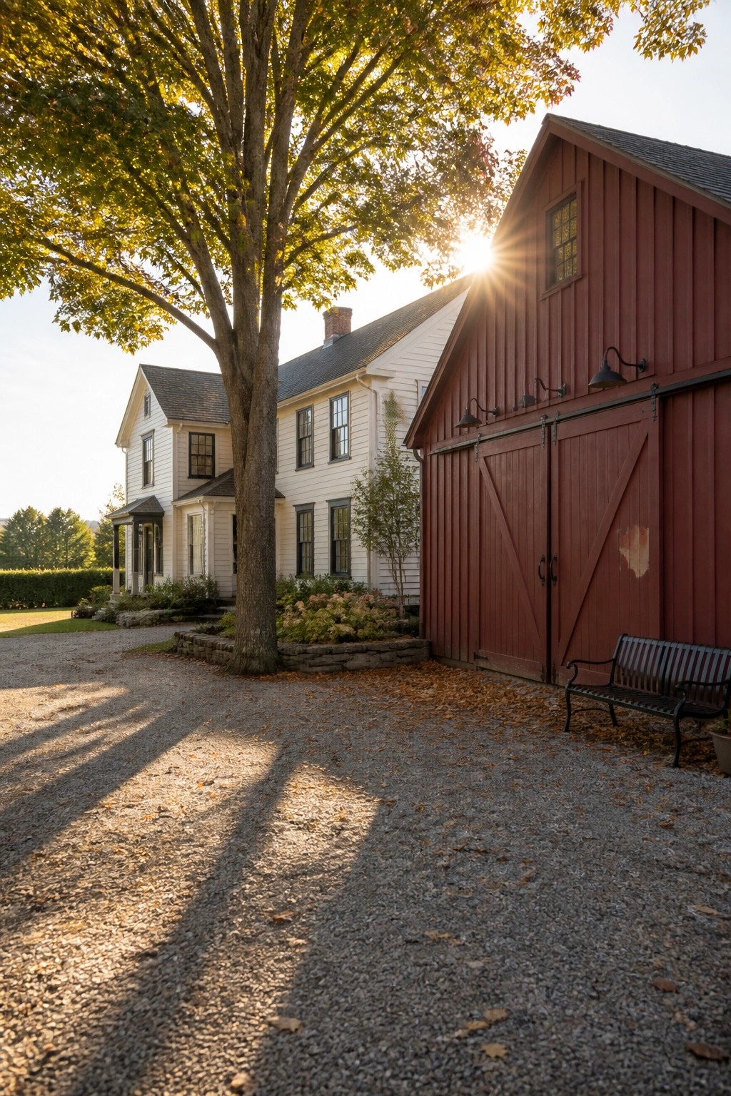 Red barn beside white house with tree casting shadows on sunny gravel driveway