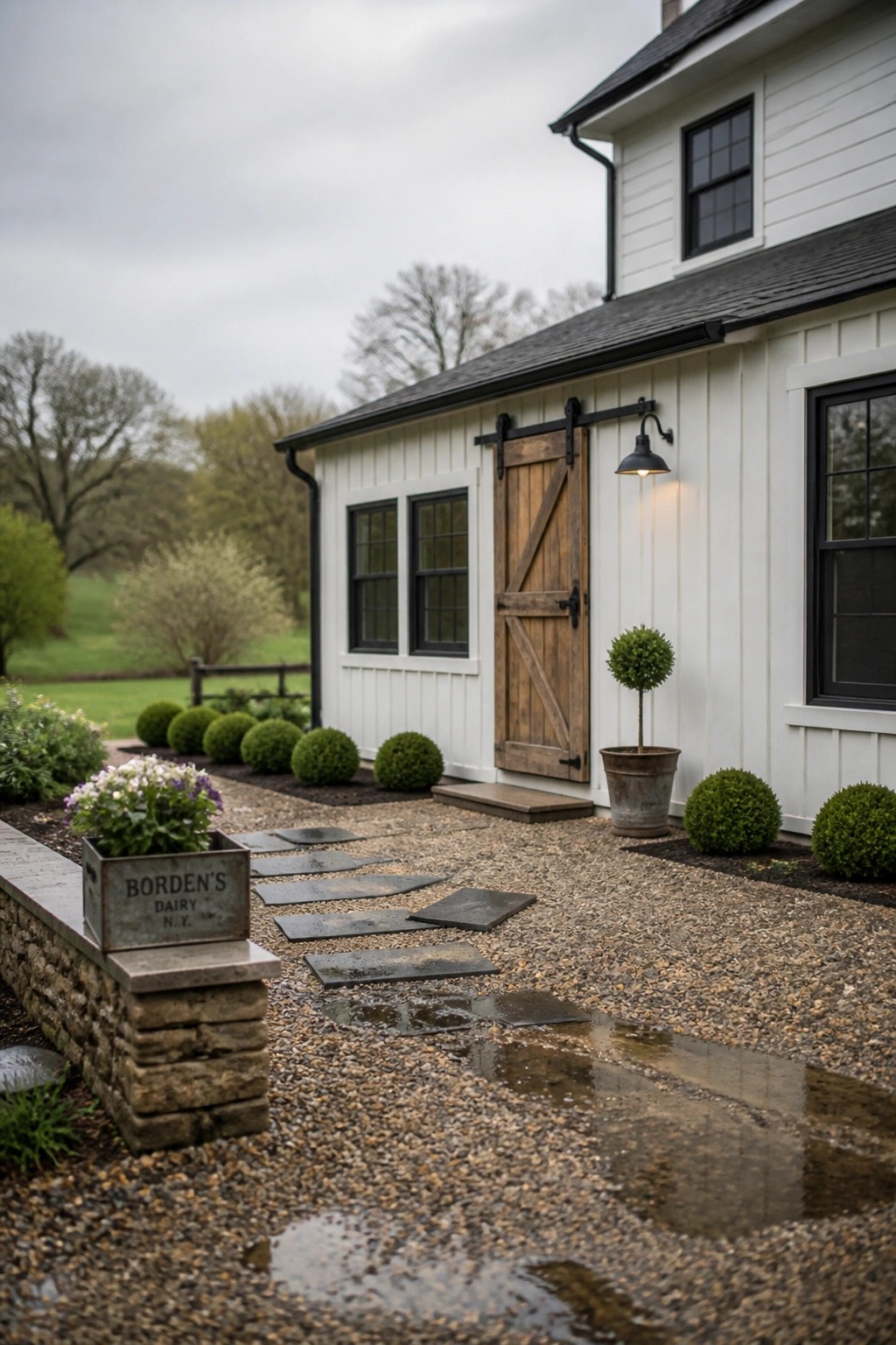 A white farmhouse exterior with a wooden barn-style door, gravel walkway lined with irregular stone pavers, and low shrubs along the edges.