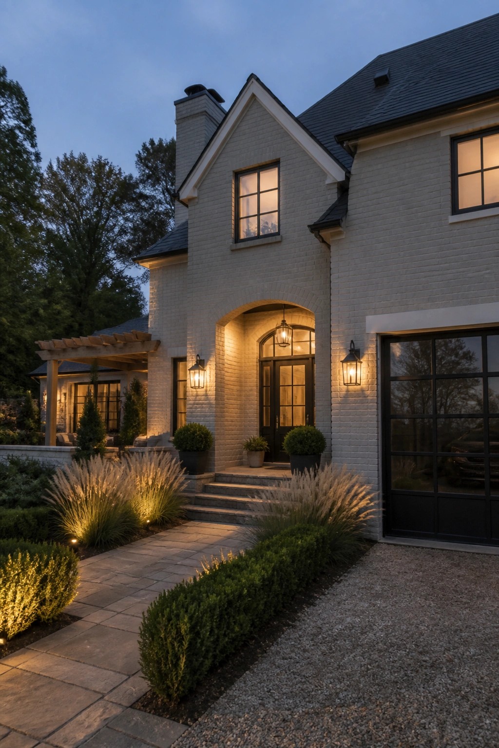 White brick house with lit arched entry, stone path, and landscaped garden at dusk.