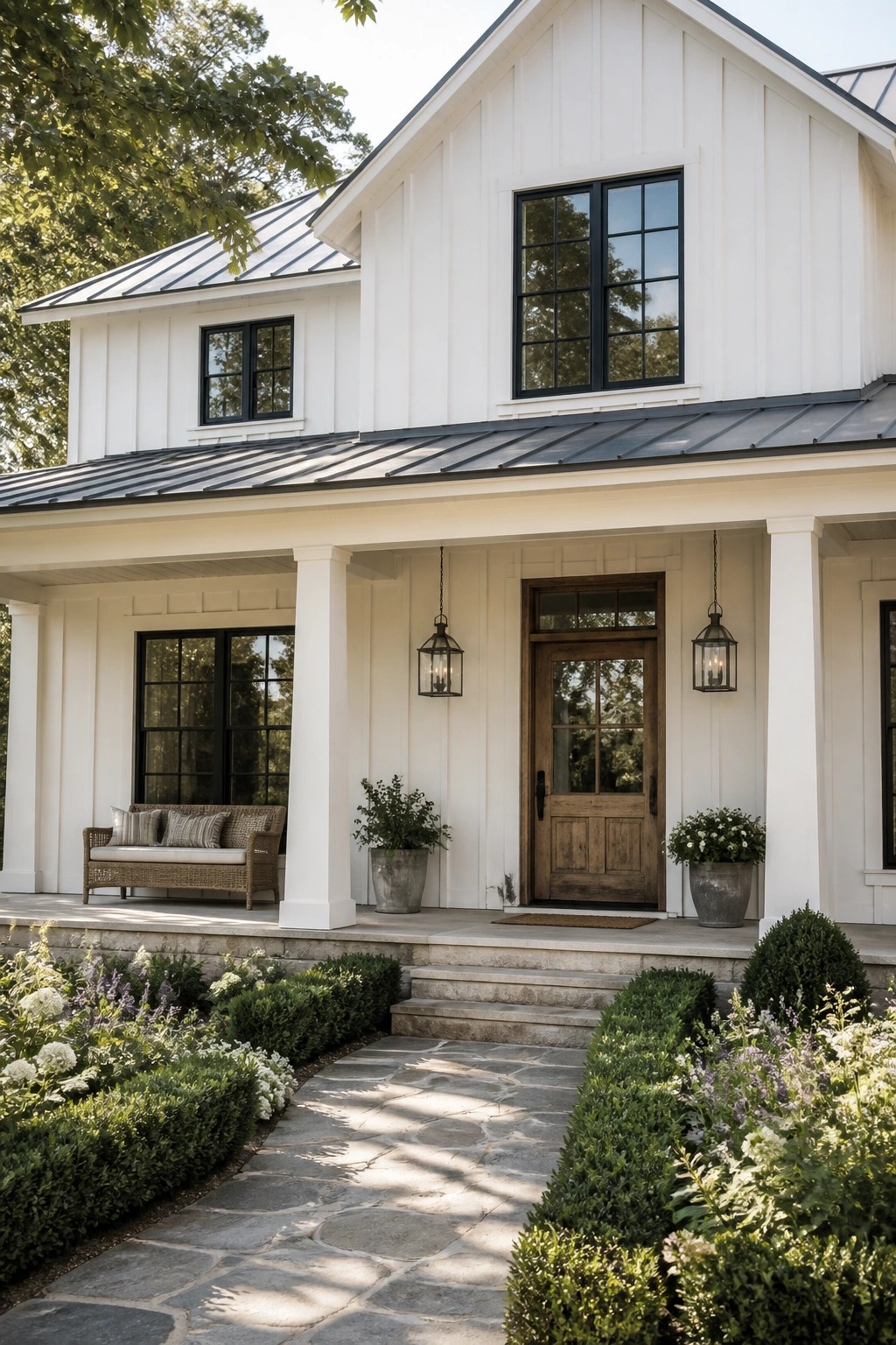 White farmhouse porch with wooden door, lanterns, wicker bench, potted plants, and stone path.