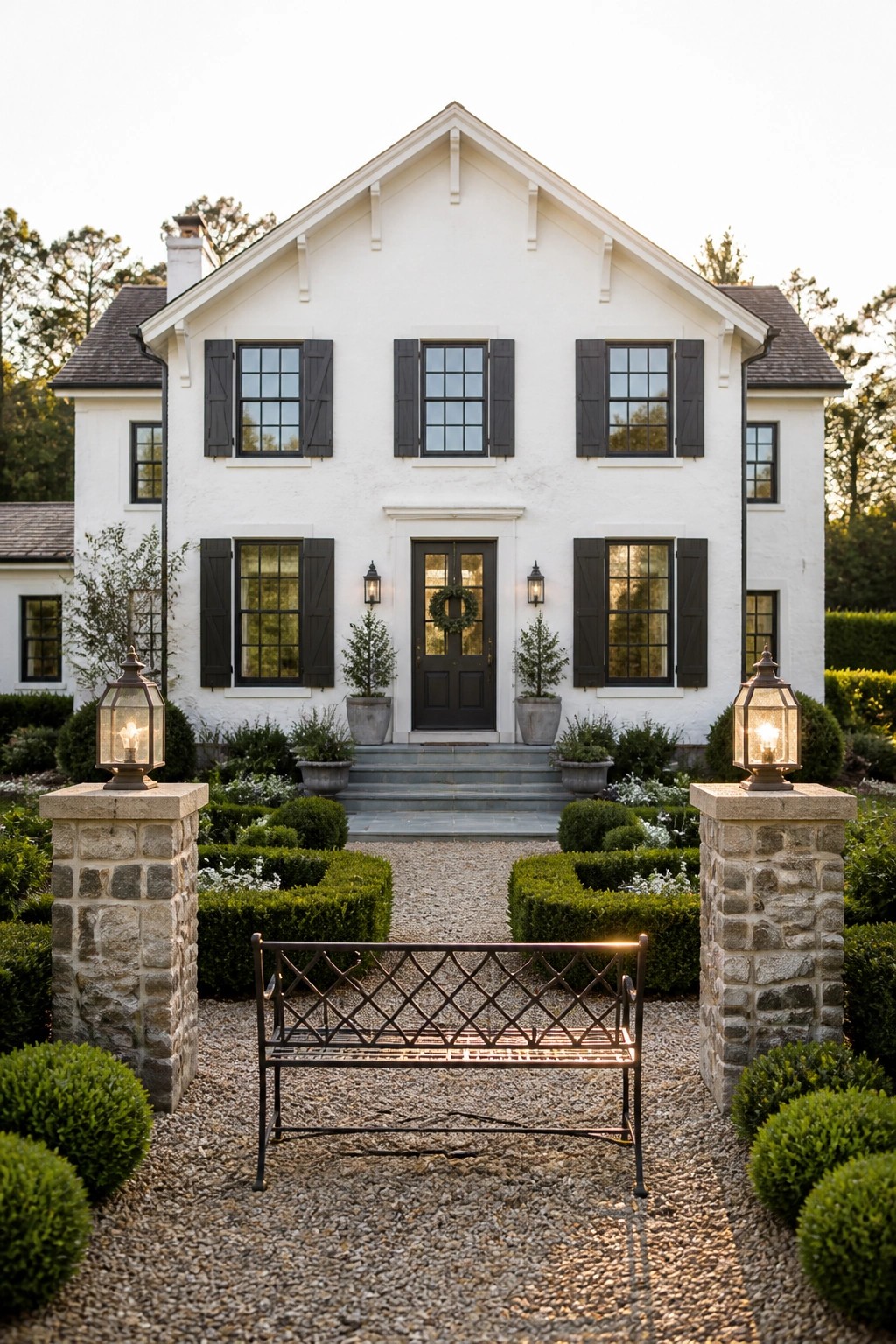 A white two-story farmhouse with black shutters, a dark front door, stone entry pillars with lanterns, and a metal bench on gravel.