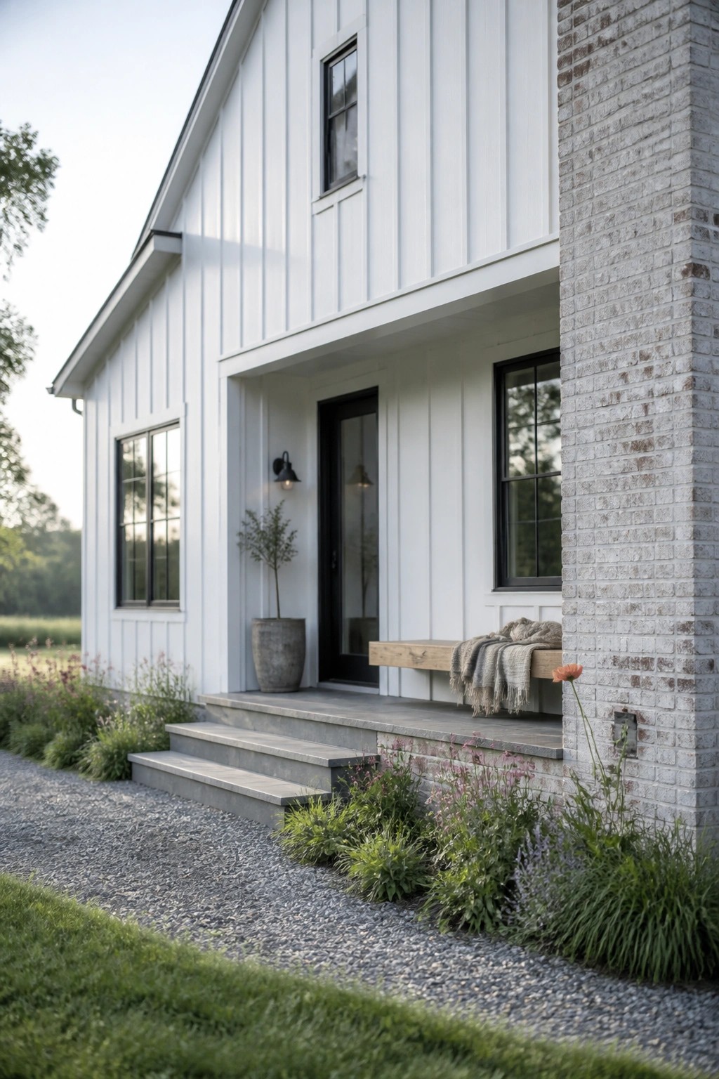 A white board and batten farmhouse exterior with black window frames, a black front door, gray brick chimney on the right, concrete steps, and low landscaping along a gravel path.