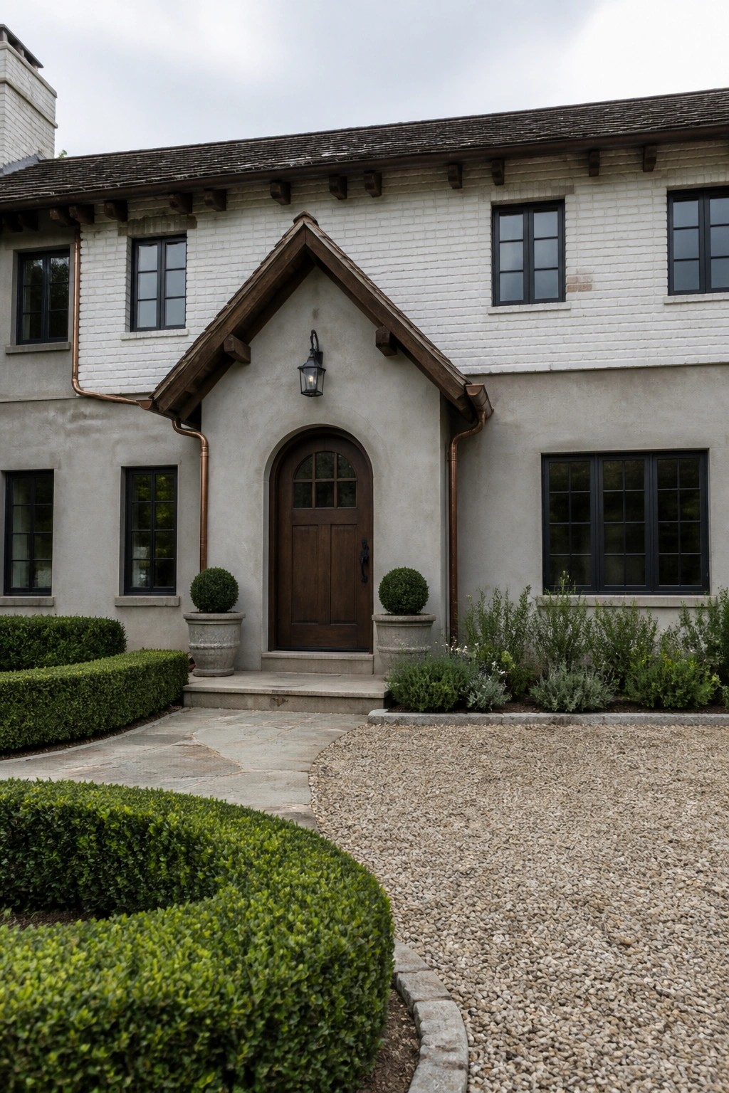 A house exterior with a wooden arched door centered under a gabled overhang, flanked by windows and potted shrubs.