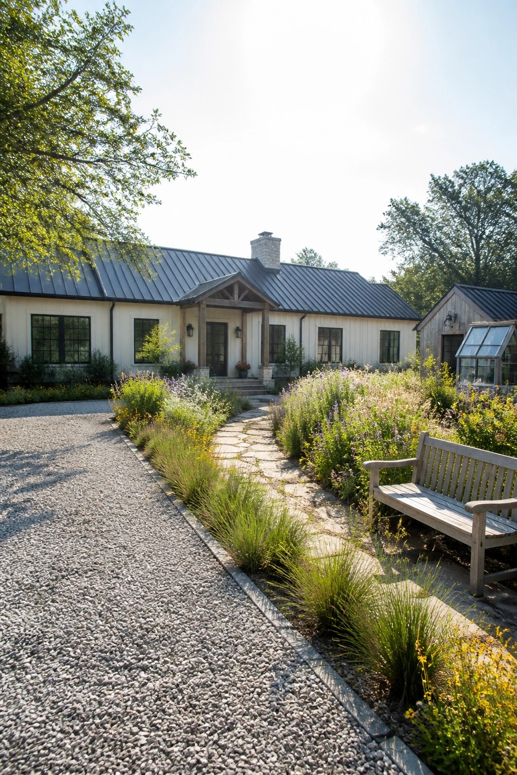A white farmhouse with dark metal roof and stone chimney has a flagstone path lined with tall grasses and wildflowers leading to the front door, next to a gravel drive and wooden bench.