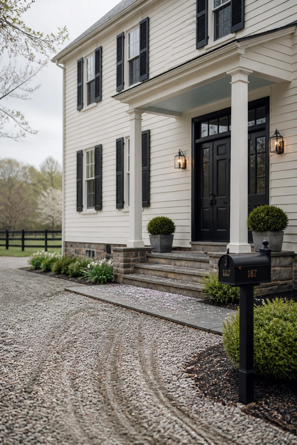 White house with black shutters and door, stone steps, mailbox by gravel drive.