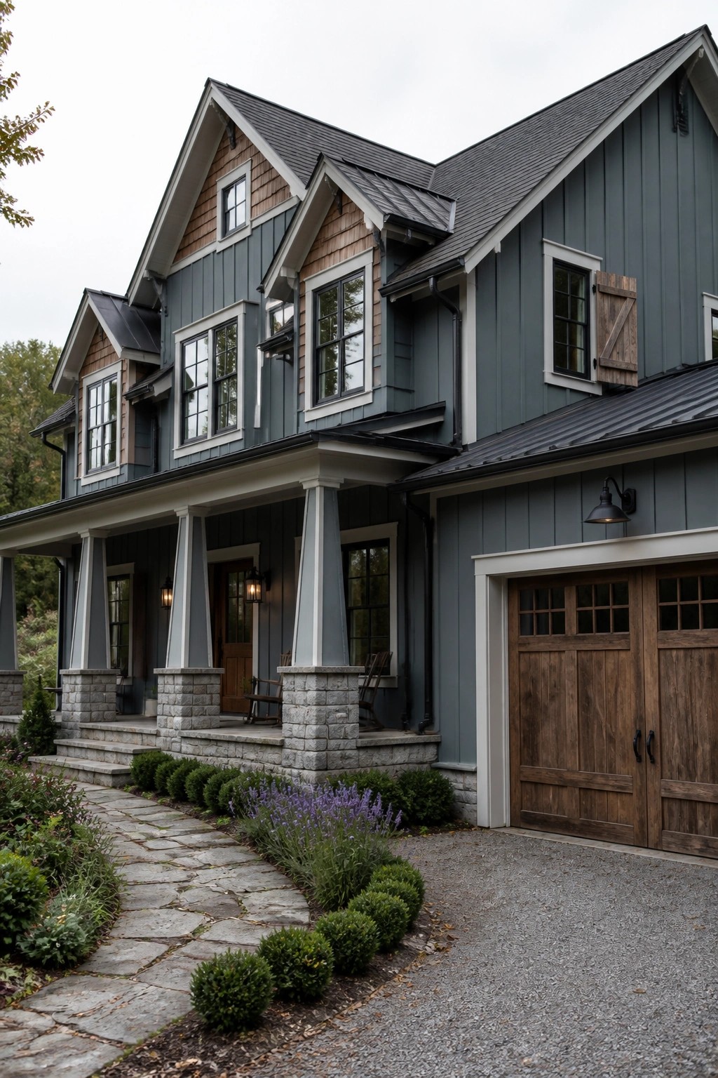 A two-story farmhouse exterior with blue-gray siding, wood accents in the gables, a stone porch, and wooden garage doors.