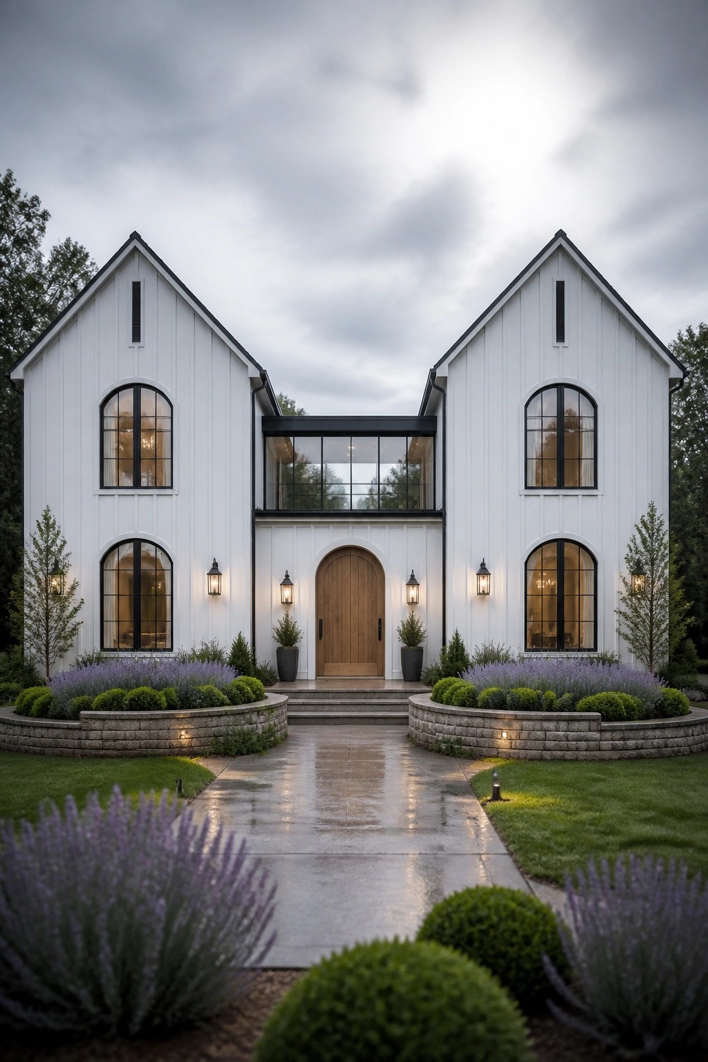 A white two-story barn-style house with two gabled wings joined by a glass bridge above a wooden arched door, flanked by arched windows and lavender landscaping in stone beds.
