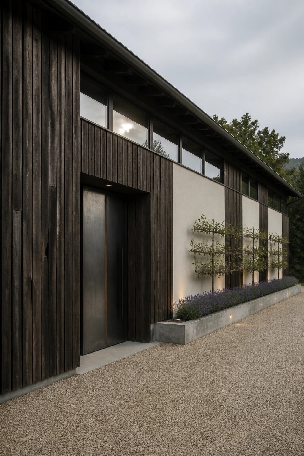 A contemporary barn house exterior showing dark vertical wood siding on the left with a dark metal door, transitioning to light walls with a row of high windows and a concrete planter bed with lavender and small trees along a gravel path.