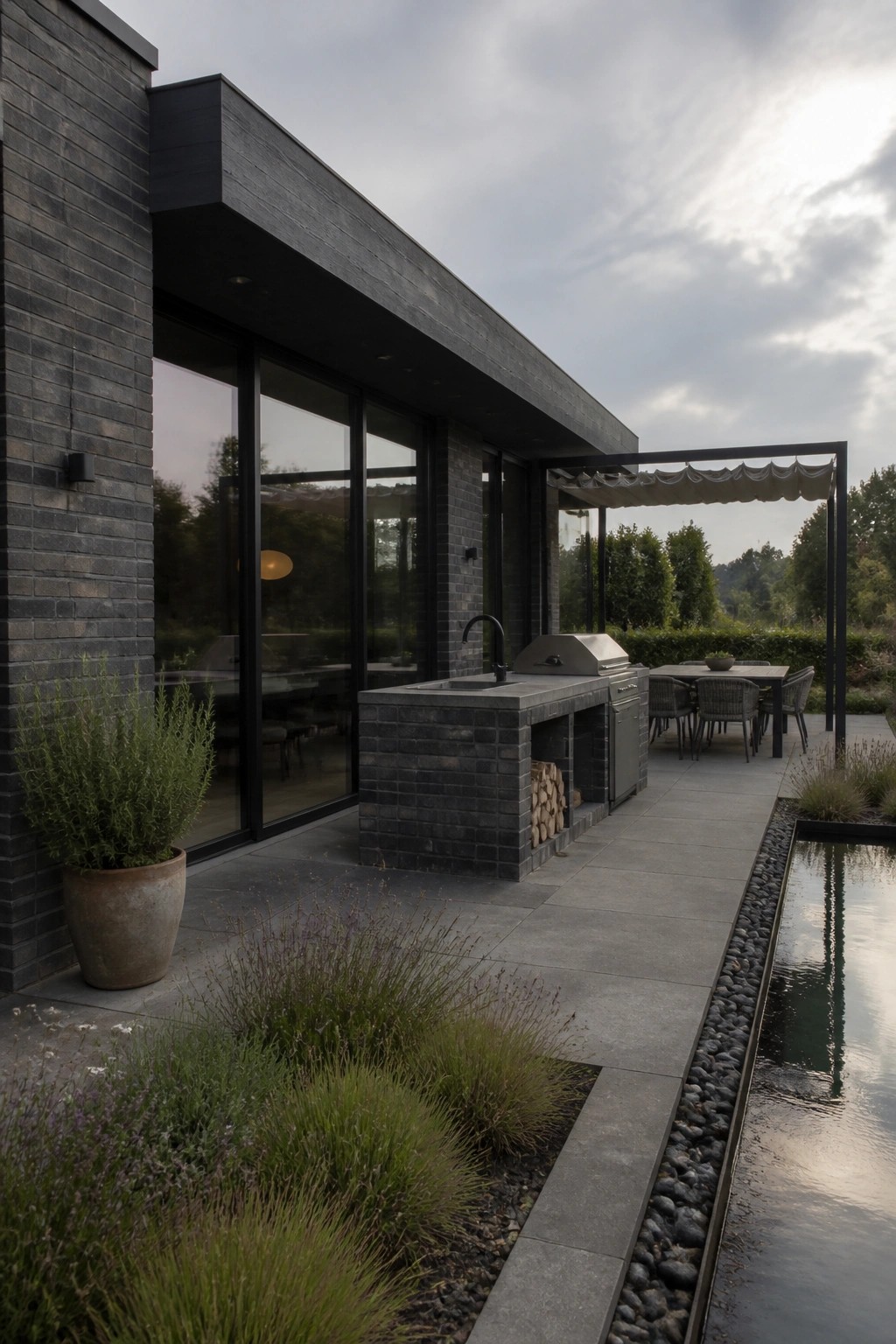 Modern dark brick house exterior with attached outdoor kitchen island, large glass sliding doors, stone patio, outdoor dining table under a pergola, and a narrow water feature along the edge.