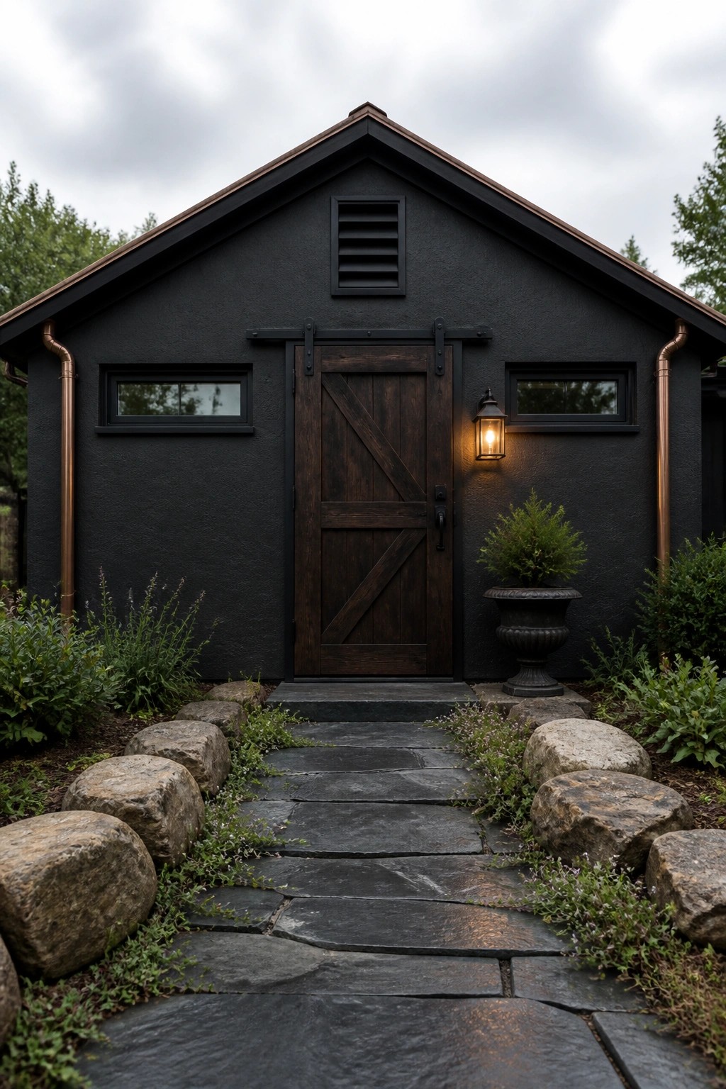A dark gray farmhouse exterior with a wooden barn door, small windows, copper downspouts, and a stone pathway leading to the entrance.
