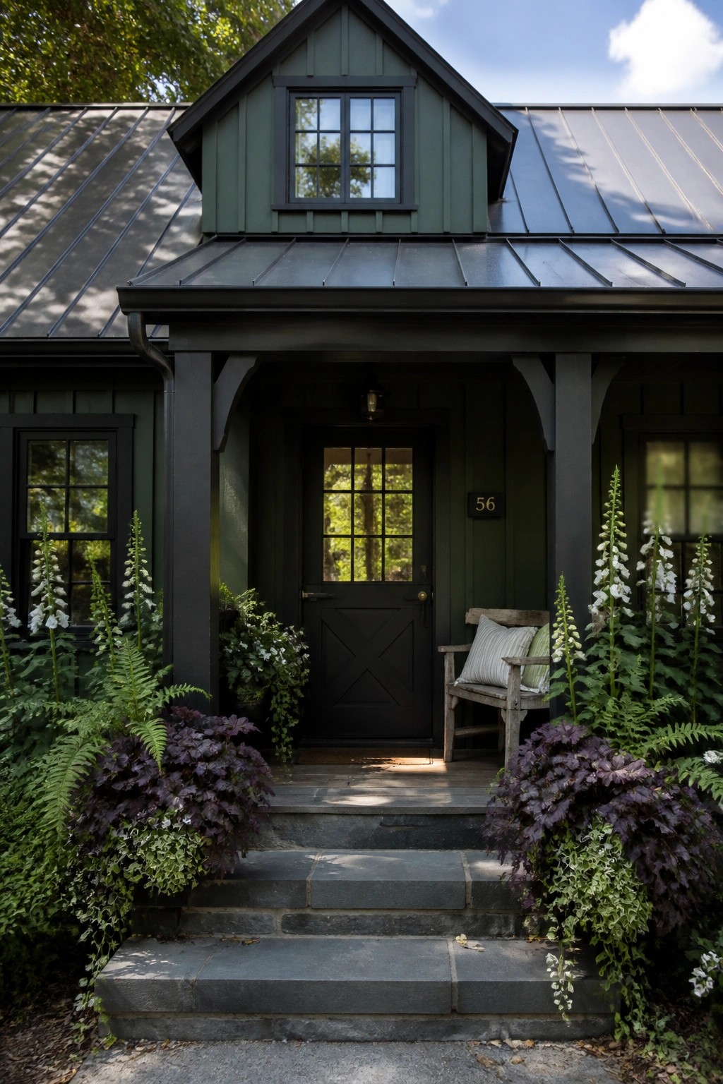 A dark green farmhouse exterior with black trim, a black front door, metal roof, and stone steps flanked by ferns and tall white flowers.