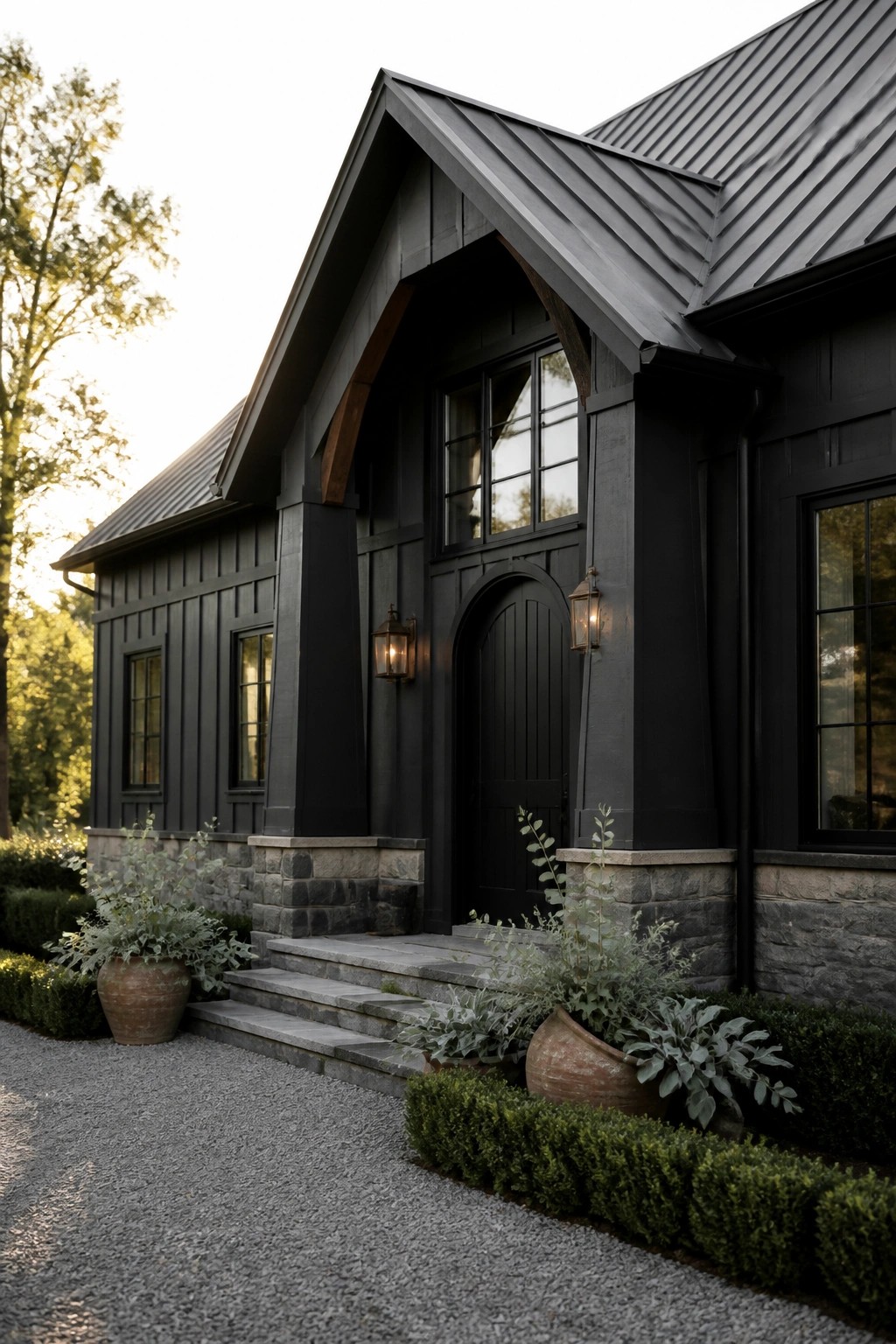 A dark painted farmhouse exterior with black siding, a metal roof, stone foundation, arched front door, and stone steps flanked by potted plants.