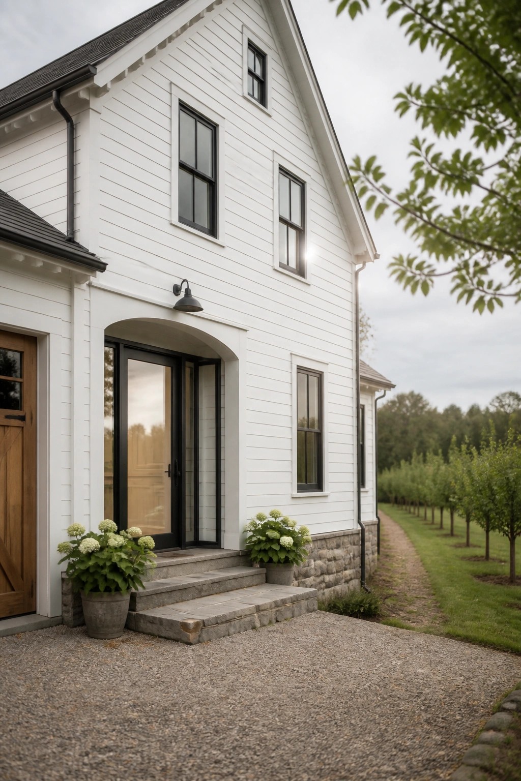 A white two-story farmhouse with black-framed windows, an arched entry door, stone foundation, and potted plants on the front steps.