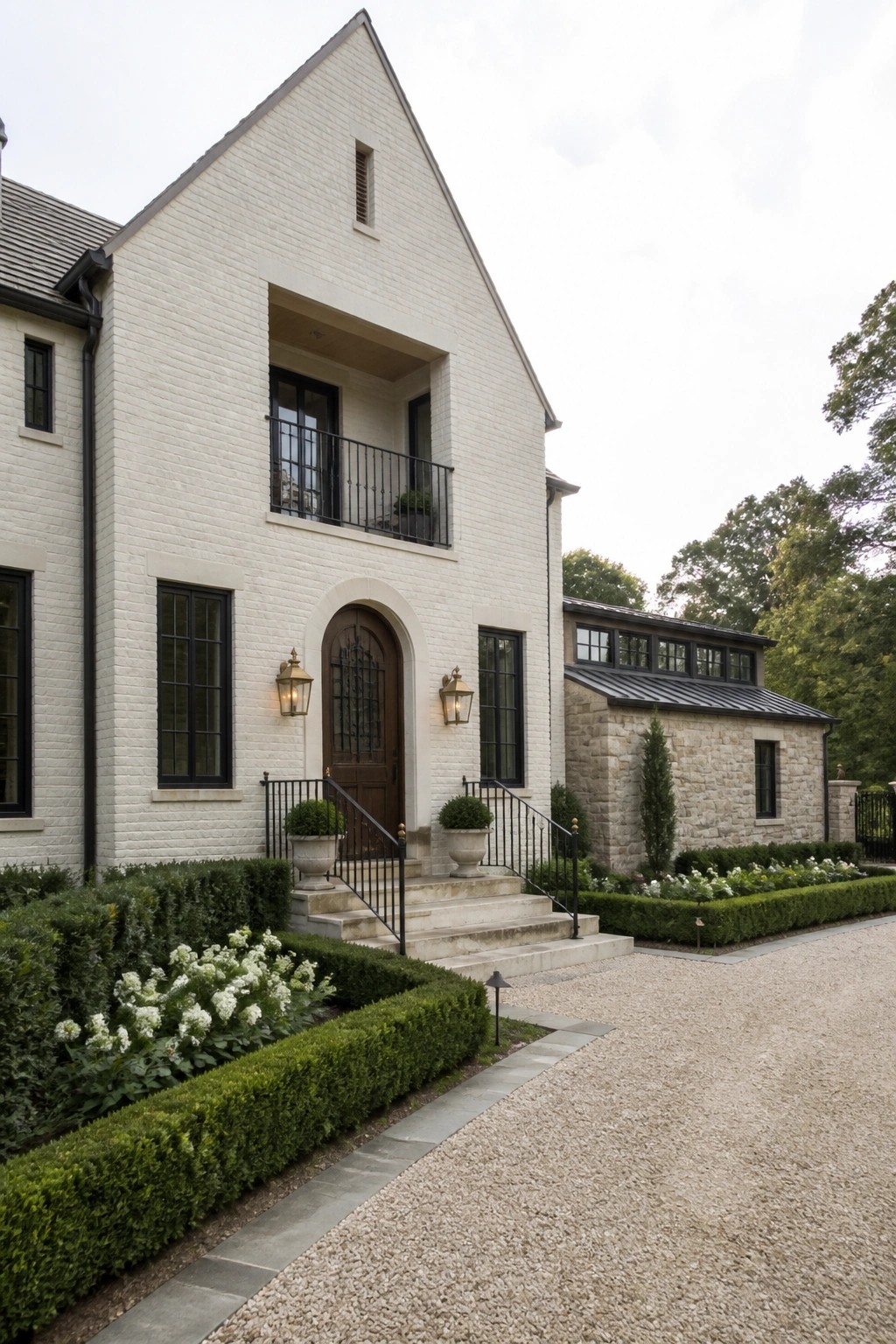White brick farmhouse exterior with an arched entry door, upper balcony, and attached stone wing on the right side.