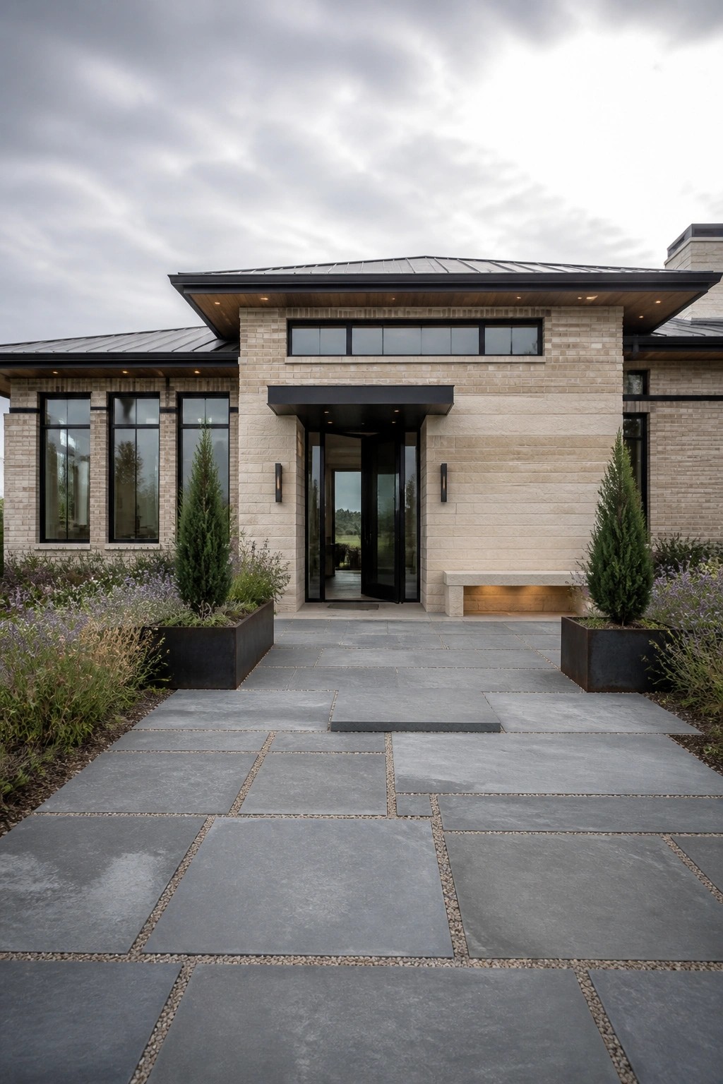 Modern stone house entrance with glass doors, gray pavers, and potted evergreens.