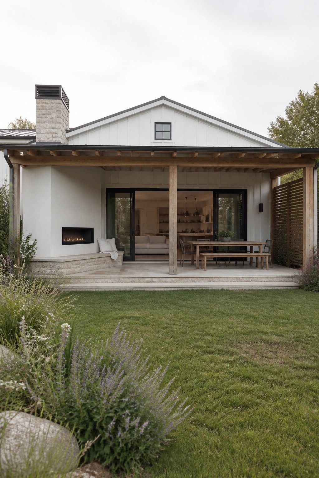 A white minimalist farmhouse exterior with a wide covered porch, wooden support beams, a stone fireplace on the left wall, and an outdoor dining table.