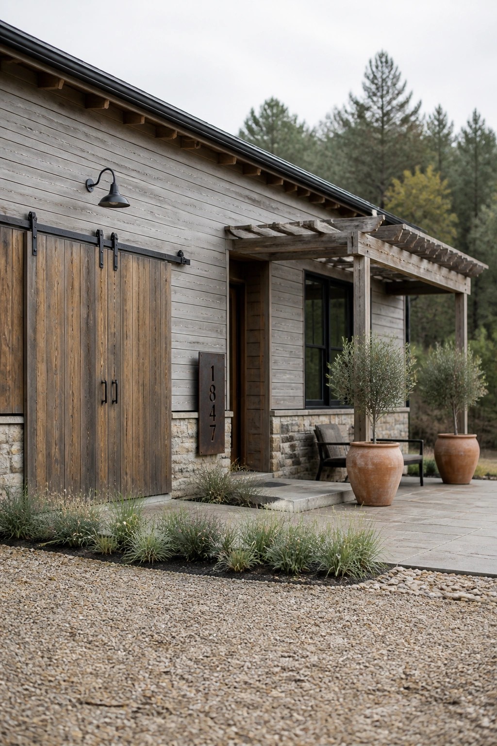 A side view of a single-story house with gray wood siding, dark sliding barn doors, a stone foundation, and a wooden pergola over the entry.
