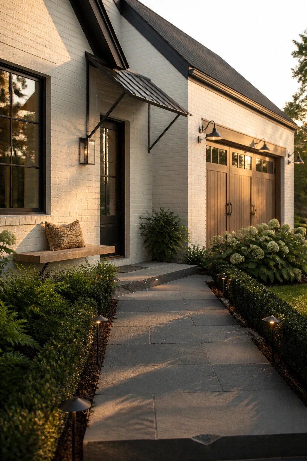 A white brick house exterior with black window frames, a black metal awning over the front door, wooden garage doors, and a stone pathway lined with low lights.