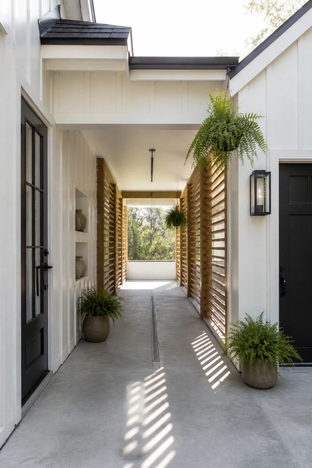 A covered concrete walkway between white buildings with black doors, vertical wood slat screens on both sides, potted ferns, and a hanging plant.