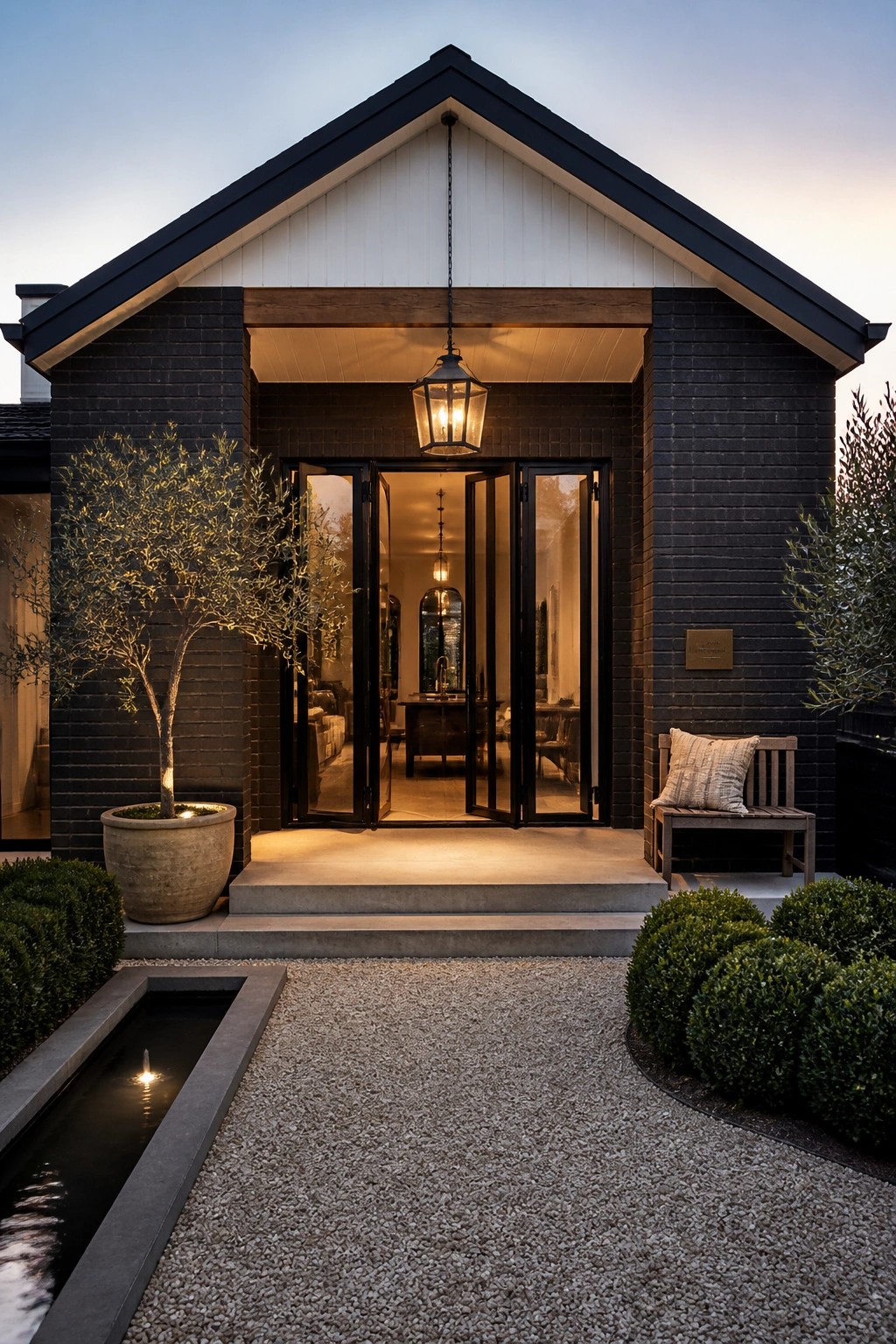 A modern farmhouse exterior features dark brick walls, a white gable, black trim around open double doors, a hanging lantern, and a gravel path with a water feature.