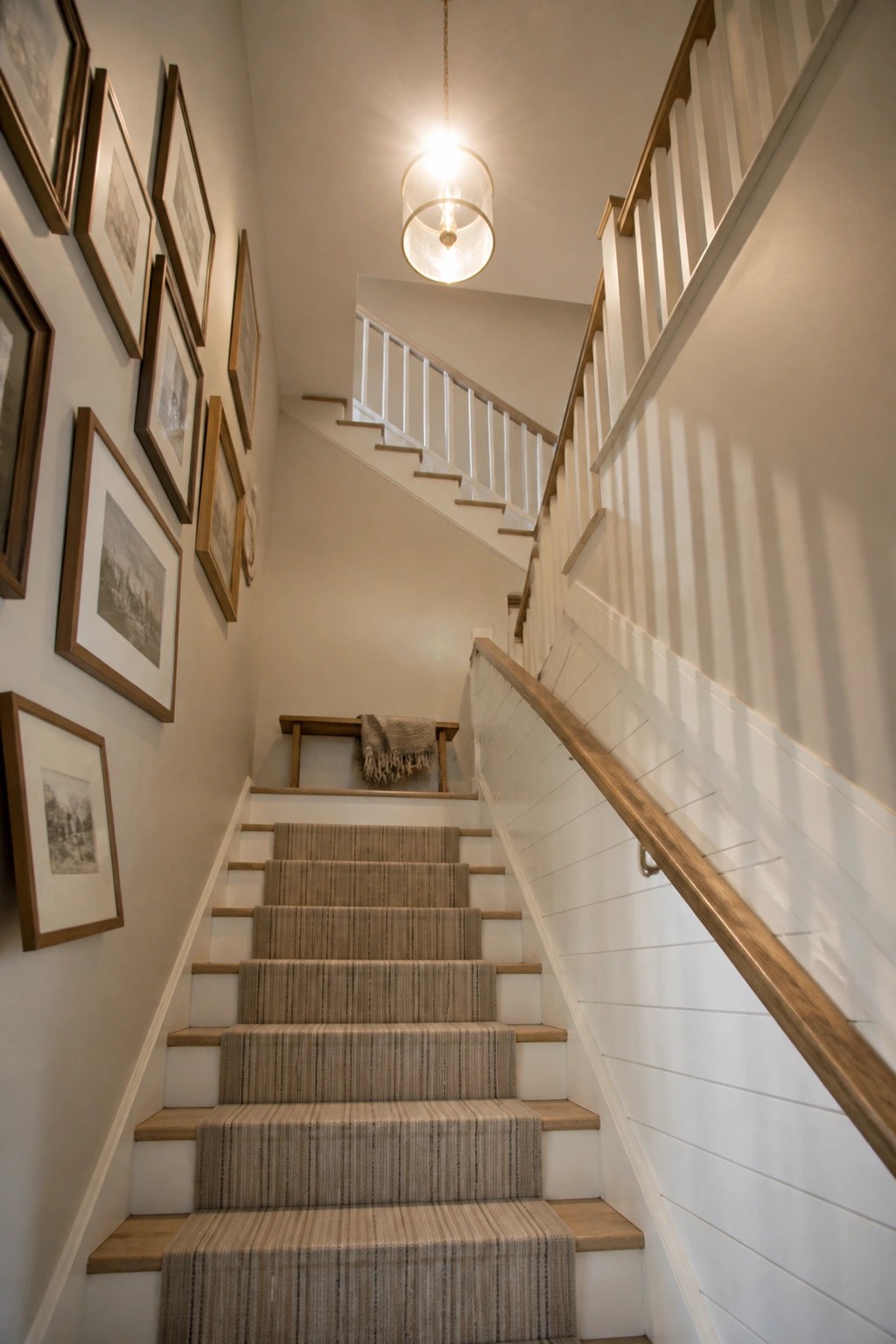 A staircase ascending with light neutral walls, white wainscoting, a wooden handrail, and framed pictures along the left wall.