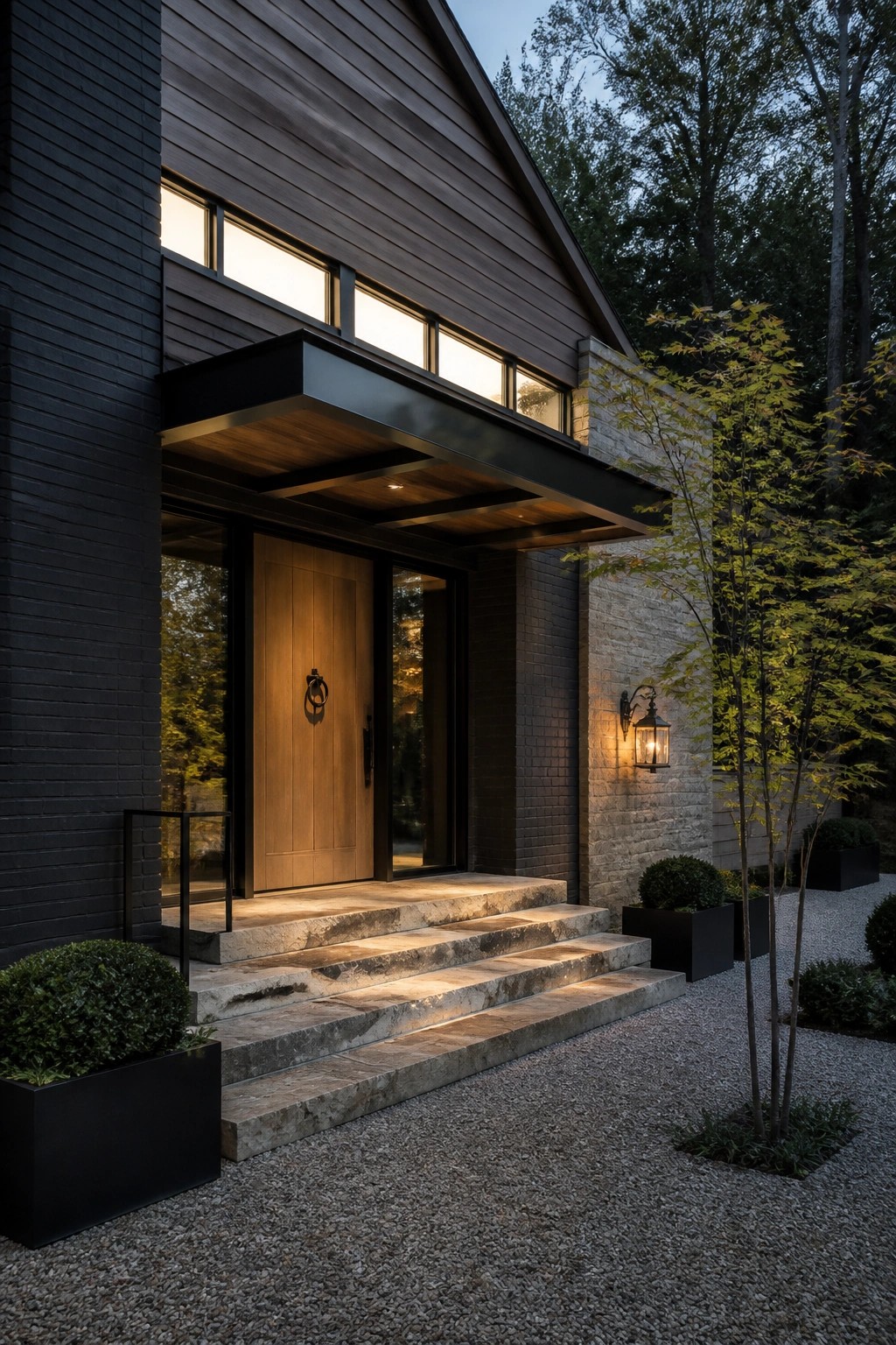 Front entrance of a modern farmhouse showing a dark metal and wood canopy above a wooden door, with stone steps and black brick siding.