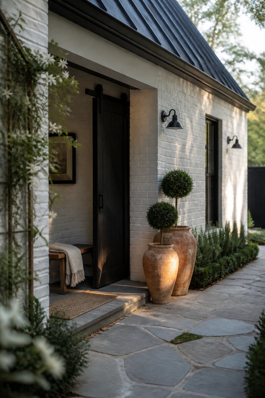 White brick exterior with a black door, black sconces, and two large terracotta pots next to a stone path.