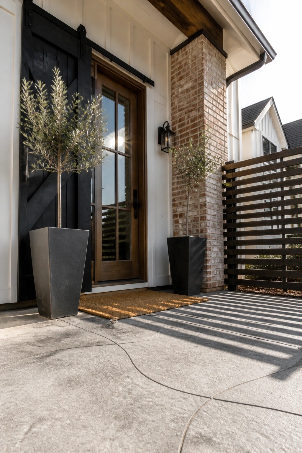A front entry with a wooden door beside a black sliding barn door, two tall black planters with trees, and a brick column on the right.
