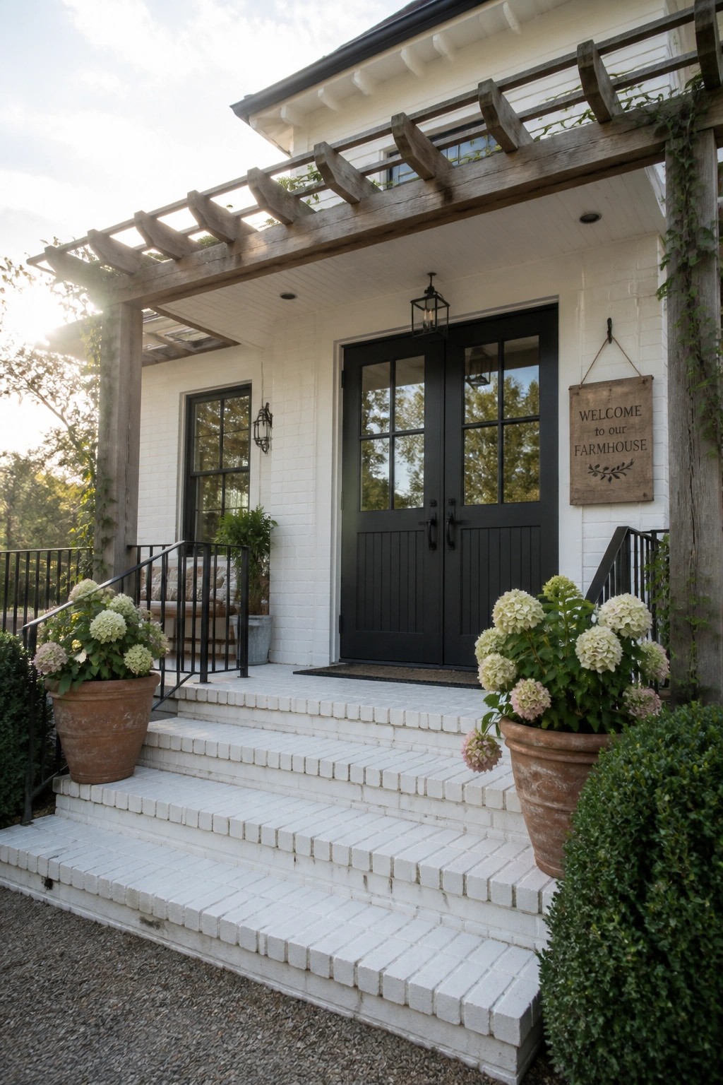 A white brick house with black double front doors under a wooden pergola, white brick steps, and potted hydrangeas on either side of the entry.