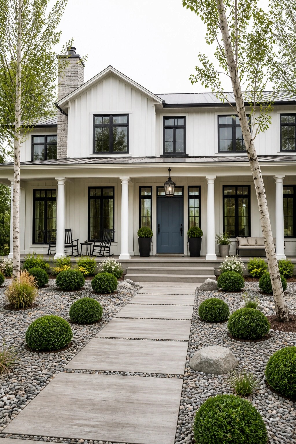 A white modern farmhouse with black window frames, a blue front door centered on a covered porch with white columns, and a gravel path with shrubs leading to the steps.