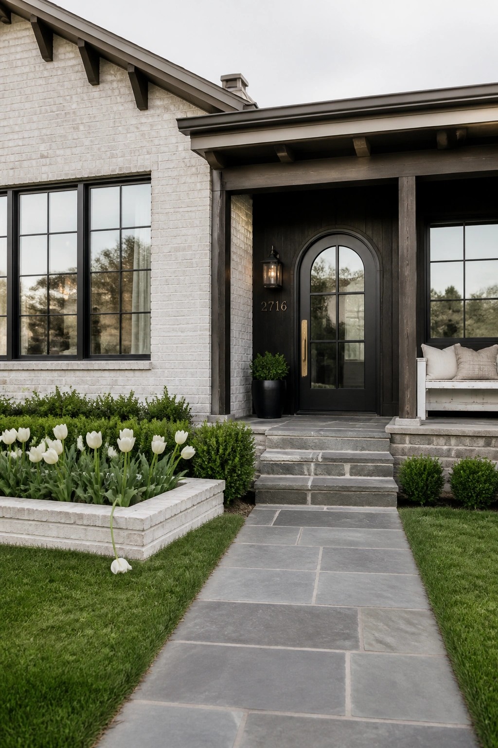 Front view of a white brick ranch house with a dark arched wood door, black-framed windows, stone steps, and a paved walkway bordered by grass and white tulips.