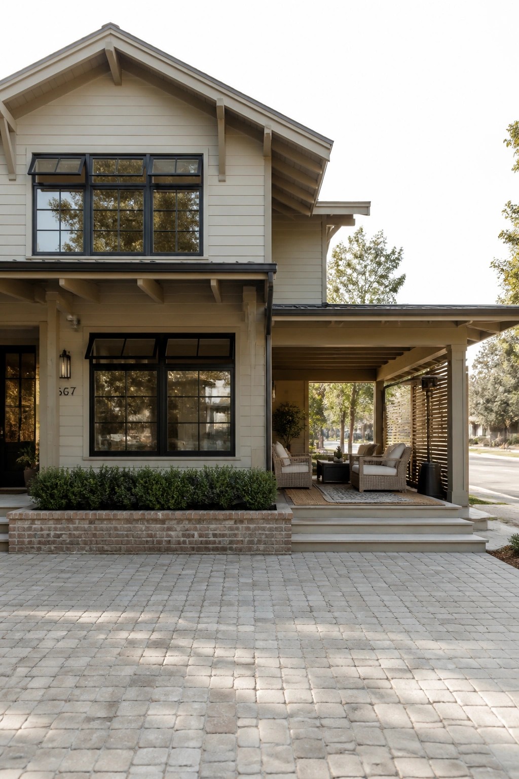 Front view of a two-story house with light siding, black-framed windows, a covered side porch with seating, a low brick planter, and a paver driveway.