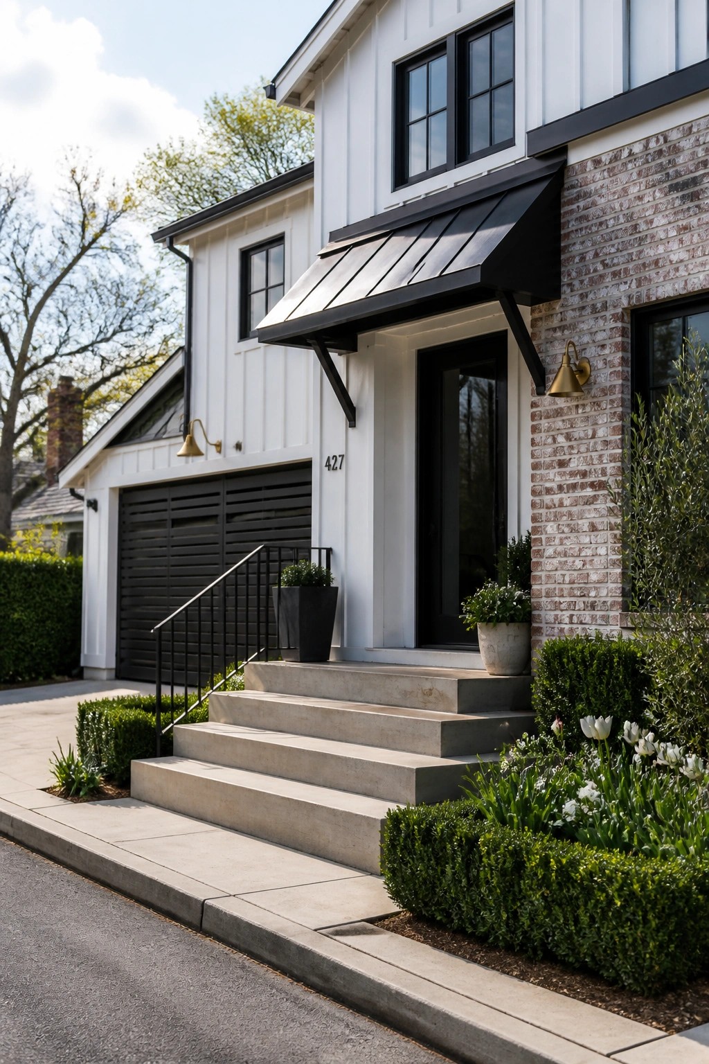 Front view of a two-story contemporary farmhouse with white vertical siding, brick on the right side, black metal awning over the entry, black door and windows, concrete steps, and front landscaping.