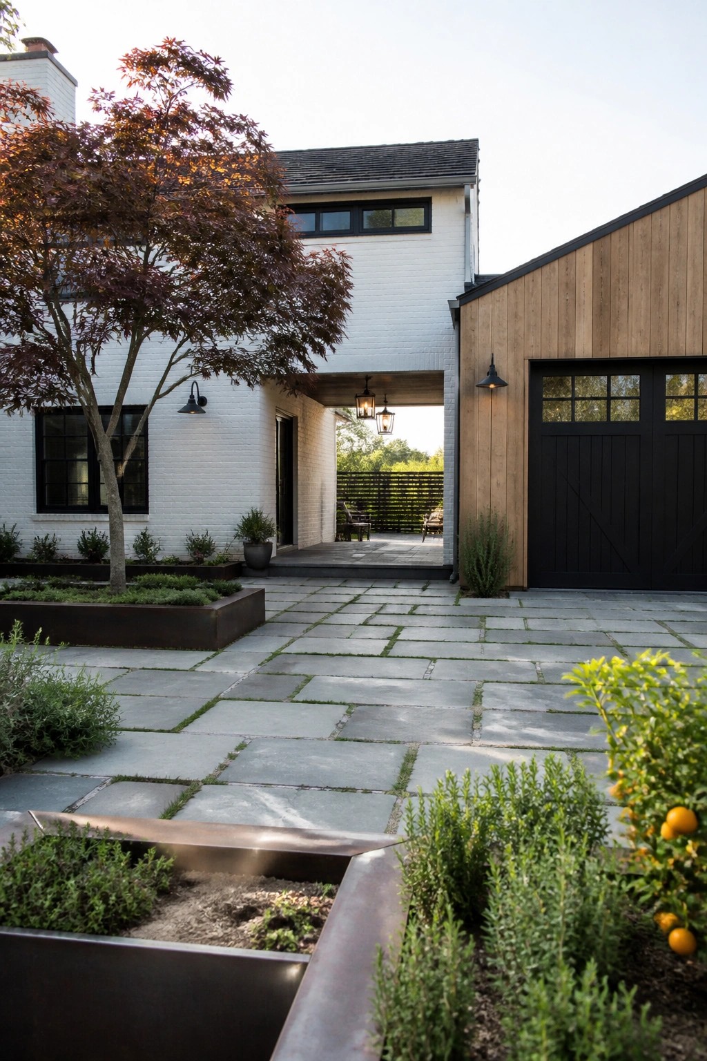 A contemporary farmhouse exterior showing white brick walls, wood siding, a black garage door, and a stone paver courtyard bordered by large dark metal planters with shrubs and trees.