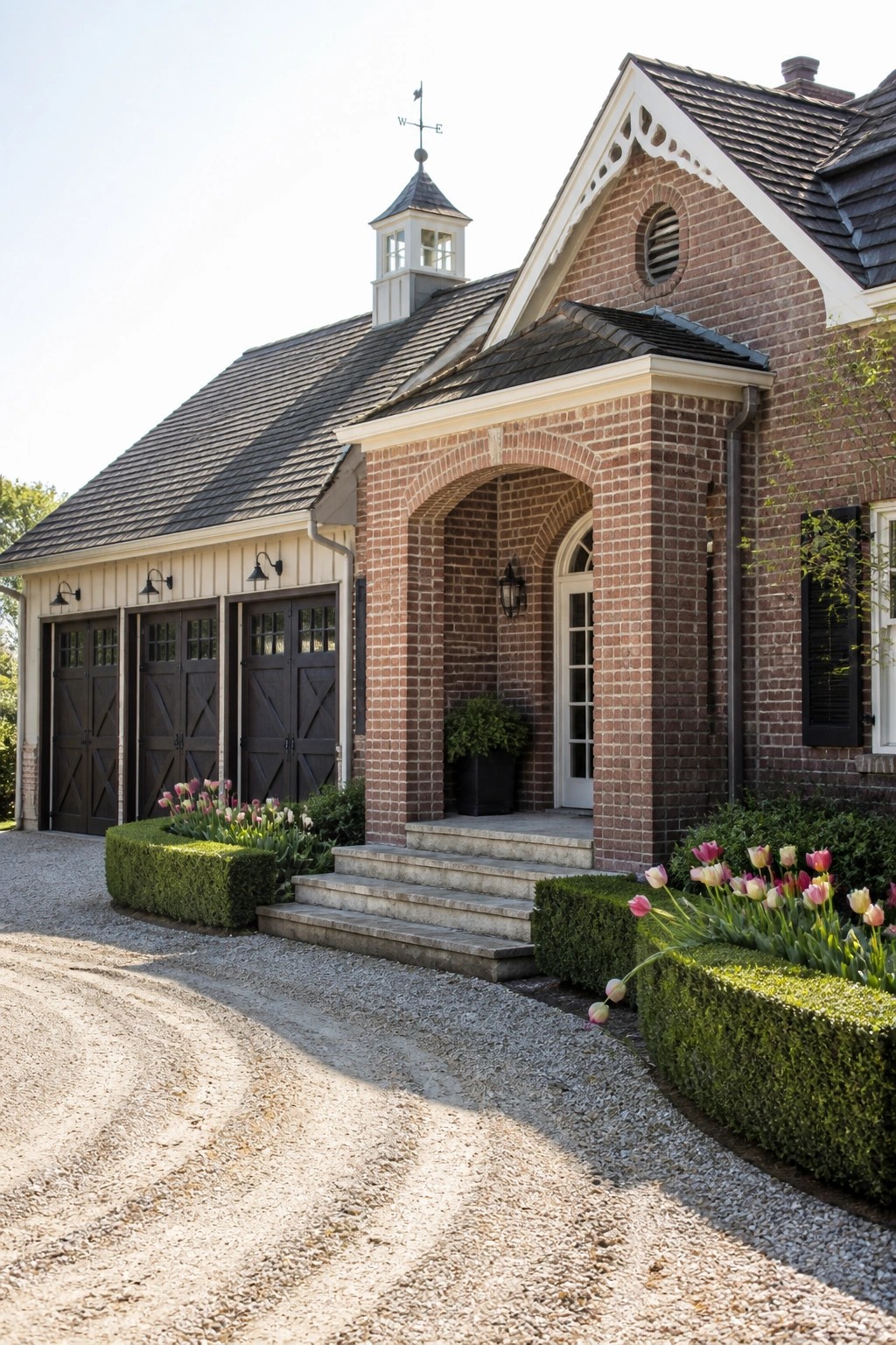 A red brick house exterior with three dark garage doors on the left, a central brick arch entry with steps, and a small cupola on the roof.
