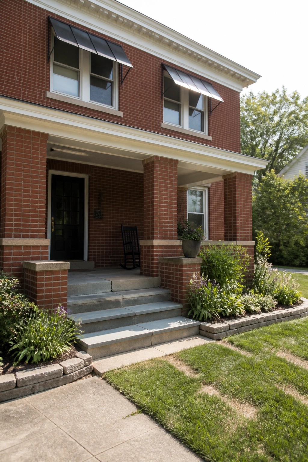 A two-story red brick house with white trim, a covered porch supported by brick columns, concrete steps, a black front door, and a small garden bed along the side.
