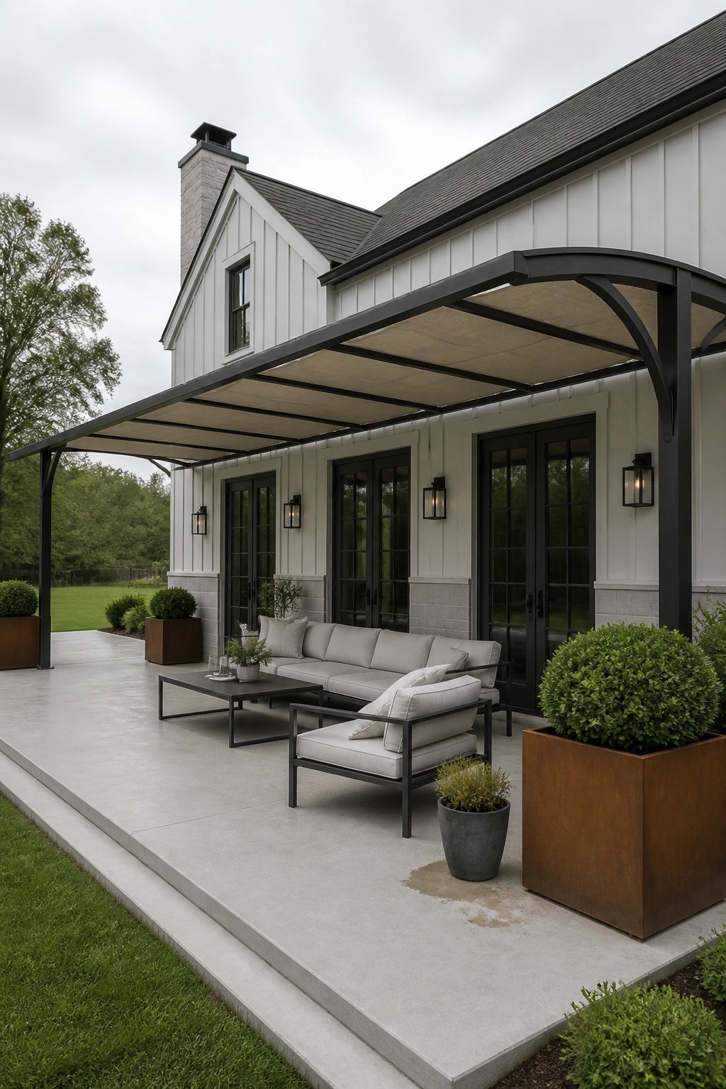 A white farmhouse exterior with black-framed doors and a covered concrete patio that includes a gray sectional sofa, two armchairs, a coffee table, and large rust-colored planters under a curved black metal awning with beige fabric.