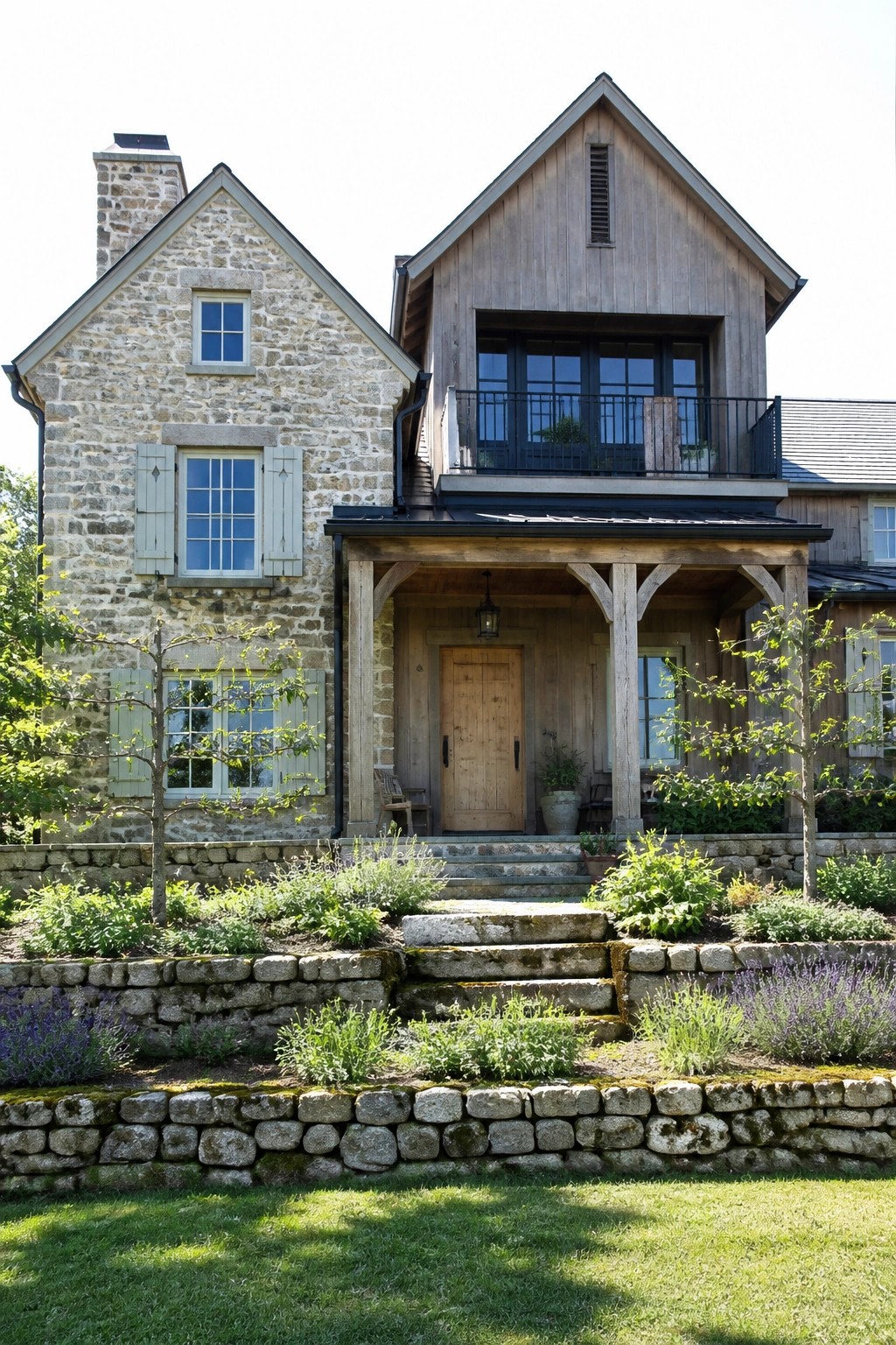 A two-story house with a stone left wing and wood right wing, a wooden front porch with steps, and low stone retaining walls with garden beds in front.